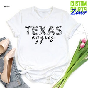 Może przedstawiać: Biała koszulka z czarno-białym wzorem kwiatowym, kt&oacute;ry tworzy napis "TEXAS aggie"
