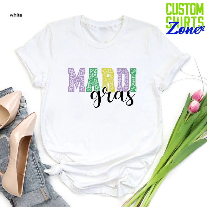 May include: White t-shirt with the text "Mardi Gras" in purple, green, and gold letters with a leopard print pattern.