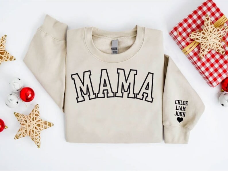 Puede incluir: Una sudadera beige con la palabra "MAMA" impresa en negro en la parte delantera. La sudadera tiene un coraz&oacute;n negro con los nombres "CHLOE, LIAM, JOHN" impresos en la manga.