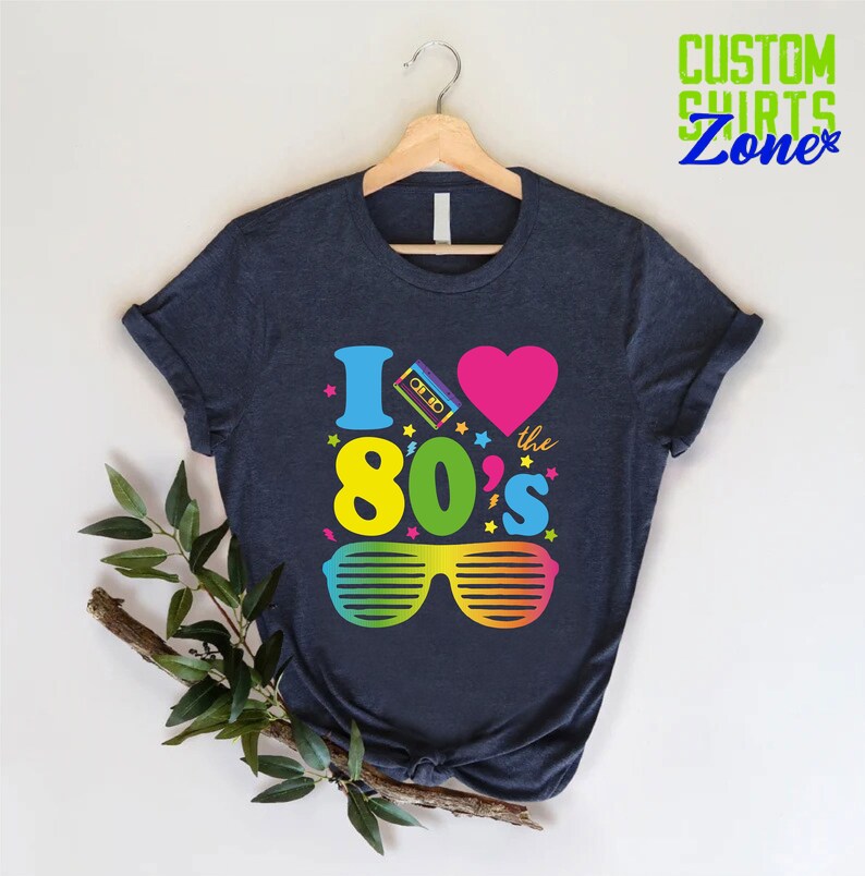 May include: A dark blue t-shirt with a graphic design that says "I love the 80's" with a pink heart, a cassette tape, and rainbow sunglasses.