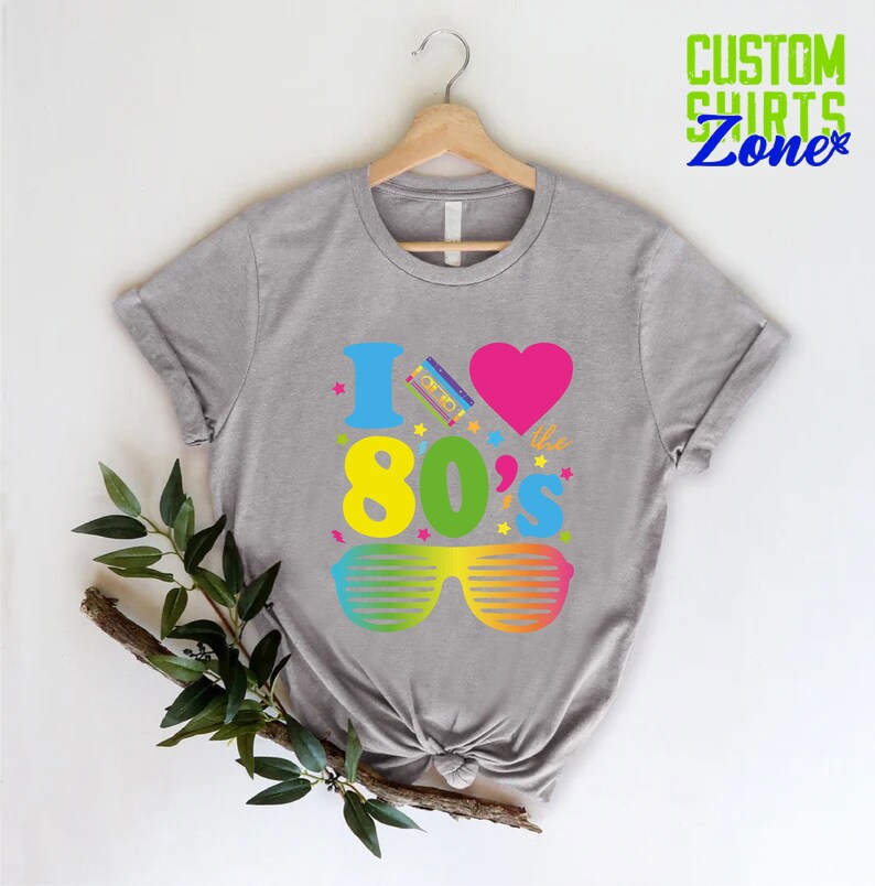 May include: Gray t-shirt with a graphic design that says "I love the 80's" with a pink heart, a cassette tape, and rainbow sunglasses.