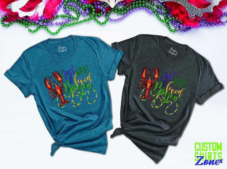 May include: Two teal and gray t-shirts with a crawfish and beads design. The text on the shirts reads "I just want to be loved in Baton Rouge".