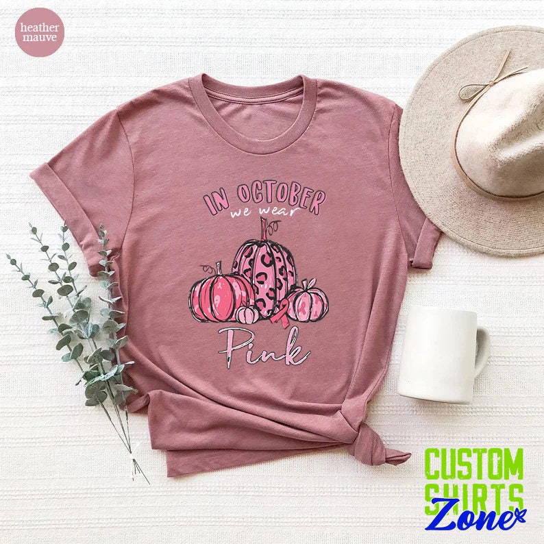 May include: A heather mauve t-shirt with the text "In October we wear Pink" and a graphic of pink pumpkins with a pink ribbon.