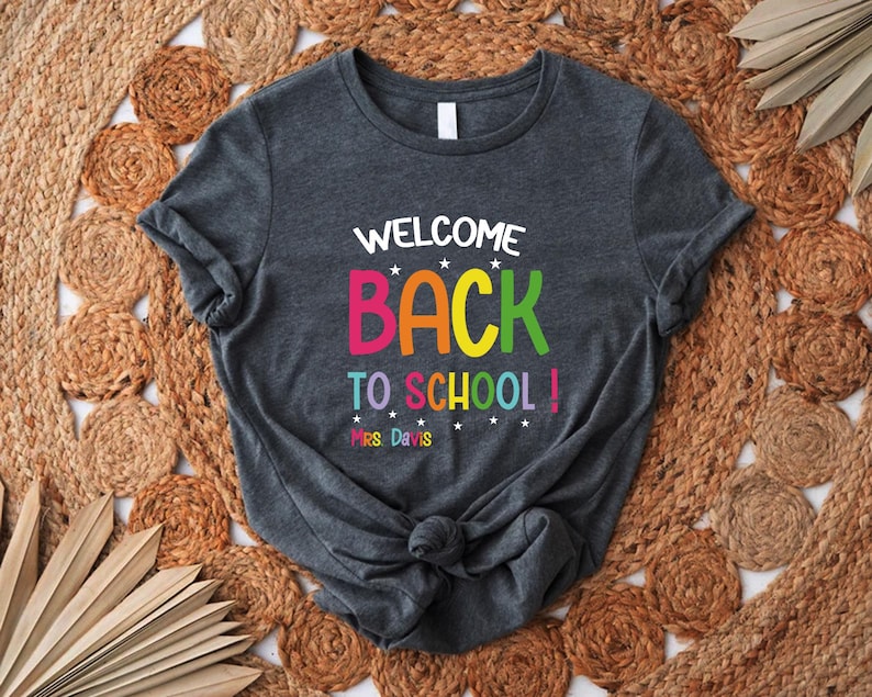 以下が含まれることがあります： カラフルな文字で「WELCOME BACK TO SCHOOL！」と書かれた濃いグレーのTシャツ。Tシャツにはテキストの下に「Mrs. Davis」という名前も含まれています。シャツは裾で結ばれています。
