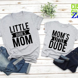 Op de afbeelding: Twee grijze t-shirts met zwarte tekst die "LITTLE DUDE'S MOM" en "MOM'S LITTLE DUDE" luidt. De shirts worden gecombineerd met opgevouwen blauwe jeans. De achtergrond is een lichte houtnerf.