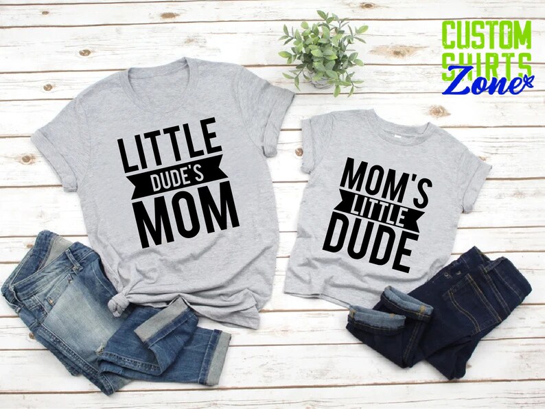 Op de afbeelding: Twee grijze t-shirts met zwarte tekst die "LITTLE DUDE'S MOM" en "MOM'S LITTLE DUDE" luidt. De shirts worden gecombineerd met opgevouwen blauwe jeans. De achtergrond is een lichte houtnerf.