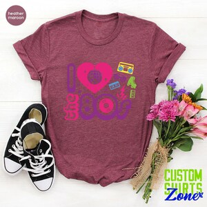 May include: A heather maroon t-shirt with a graphic design that says "I love the 80s" with a heart and 80s icons like a cassette tape, roller skates, and a record.