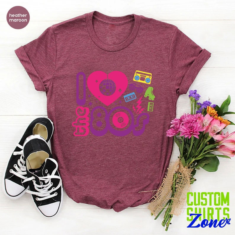 May include: A heather maroon t-shirt with a graphic design that says "I love the 80s" with a heart and 80s icons like a cassette tape, roller skates, and a record.