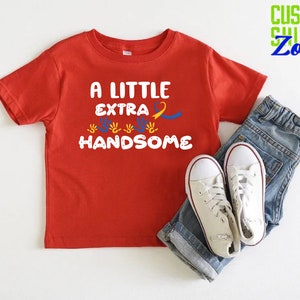 Pode incluir: Camiseta vermelha de manga curta com o texto "A Little Extra Handsome" em letras azuis e brancas. A camiseta apresenta uma fita amarela e azul e v&aacute;rias impress&otilde;es de m&atilde;os azuis.