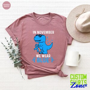 May include: Heather mauve t-shirt featuring a blue dinosaur wearing sunglasses and a blue ribbon. The text reads "IN NOVEMBER WE WEAR BLUE" with blue awareness ribbons. The shirt is a casual wear item.