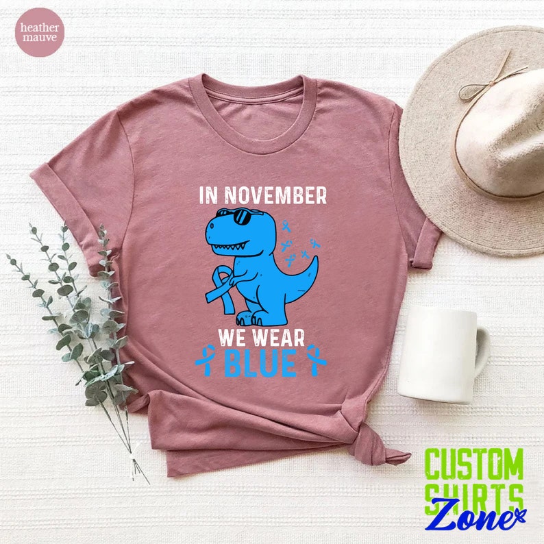 May include: Heather mauve t-shirt featuring a blue dinosaur wearing sunglasses and a blue ribbon. The text reads "IN NOVEMBER WE WEAR BLUE" with blue awareness ribbons. The shirt is a casual wear item.