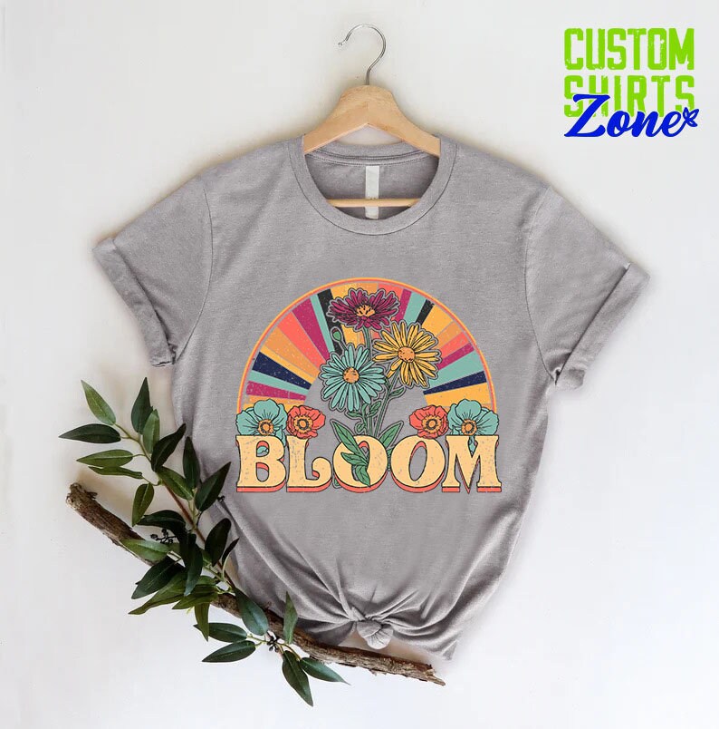 May include: A gray t-shirt with a retro-style graphic of flowers and a rainbow arc. The text "BLOOM" is printed in a vintage font below the graphic.