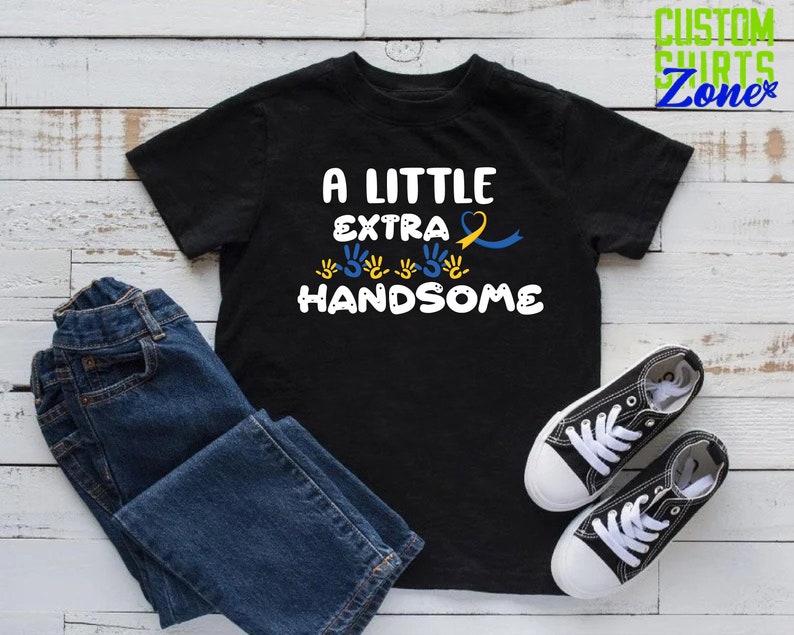 Pode incluir: Camiseta preta com o texto "A Little Extra Handsome" em branco. O texto &eacute; rodeado por impress&otilde;es de m&atilde;os amarelas e azuis e uma fita amarela e azul.