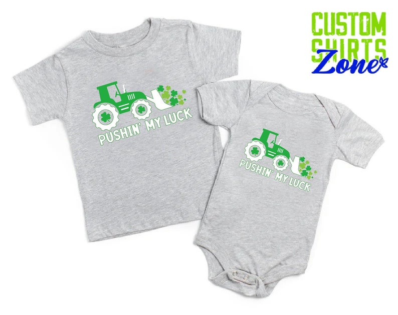 May include: Two gray shirts with a green tractor and shamrocks. The larger shirt says "Pushin' My Luck" and the smaller shirt says "Pushin' My Luck".