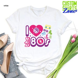 May include: White t-shirt with a pink heart and the text "I love the 80s" in purple and pink. The heart has a disco ball inside. There are other 80s icons around the text, including roller skates, sunglasses, a cassette tape, and a record.