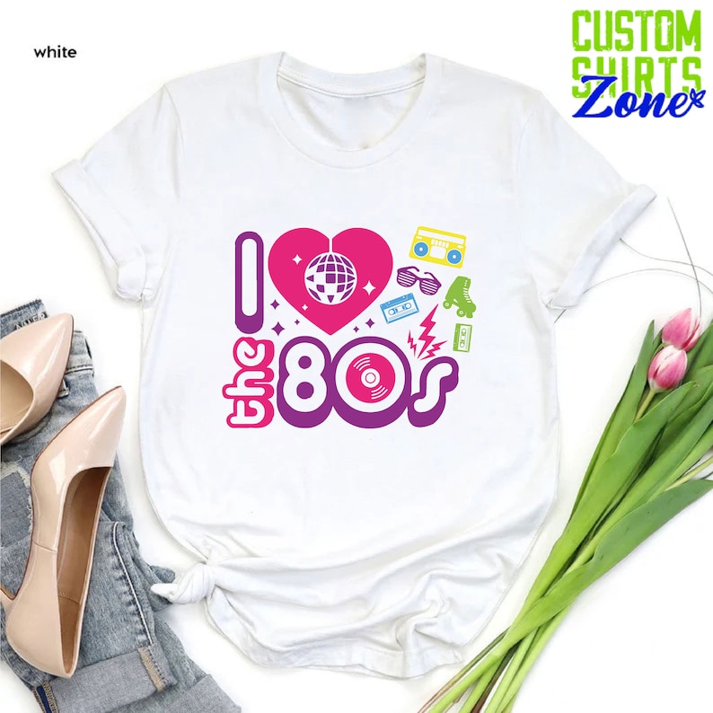 May include: White t-shirt with a pink heart and the text "I love the 80s" in purple and pink. The heart has a disco ball inside. There are other 80s icons around the text, including roller skates, sunglasses, a cassette tape, and a record.