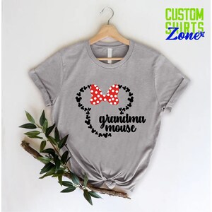 Op de afbeelding: Een grijs t-shirt met een Minnie Mouse-ontwerp. Het ontwerp bevat een rode strik met witte stippen, zwarte muizenoren en de woorden "grandma mouse" in zwart schrift. Het t-shirt hangt aan een houten hanger.