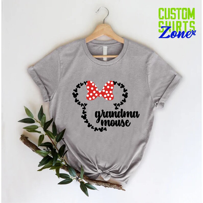 Op de afbeelding: Een grijs t-shirt met een Minnie Mouse-ontwerp. Het ontwerp bevat een rode strik met witte stippen, zwarte muizenoren en de woorden "grandma mouse" in zwart schrift. Het t-shirt hangt aan een houten hanger.