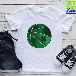 Puede incluir: Camiseta blanca con un dise&ntilde;o de baloncesto verde y un tr&eacute;bol de cuatro hojas. El baloncesto tiene costuras blancas y un tr&eacute;bol verde.