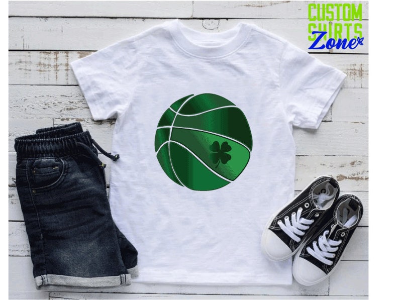 Puede incluir: Camiseta blanca con un dise&ntilde;o de baloncesto verde y un tr&eacute;bol de cuatro hojas. El baloncesto tiene costuras blancas y un tr&eacute;bol verde.