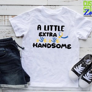 Pode incluir: Camiseta branca com o texto "A Little Extra Handsome" em preto. O texto &eacute; rodeado por impress&otilde;es de m&atilde;os amarelas e uma fita azul e amarela.
