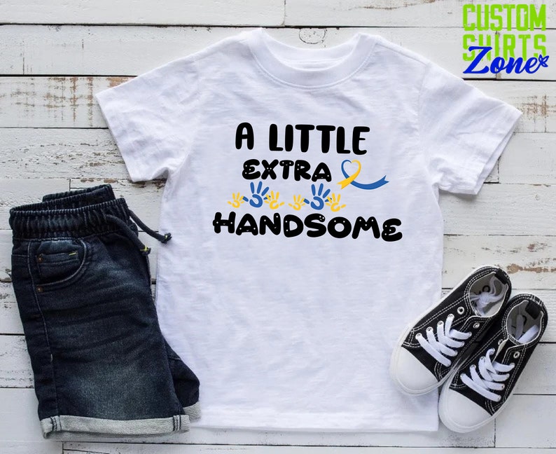 Pode incluir: Camiseta branca com o texto "A Little Extra Handsome" em preto. O texto &eacute; rodeado por impress&otilde;es de m&atilde;os amarelas e uma fita azul e amarela.