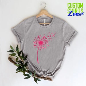 Op de afbeelding: Grijs T-shirt met een roze paardenbloemontwerp gemaakt van roze linten. De paardenbloem waait in de wind. Het shirt hangt aan een hanger.
