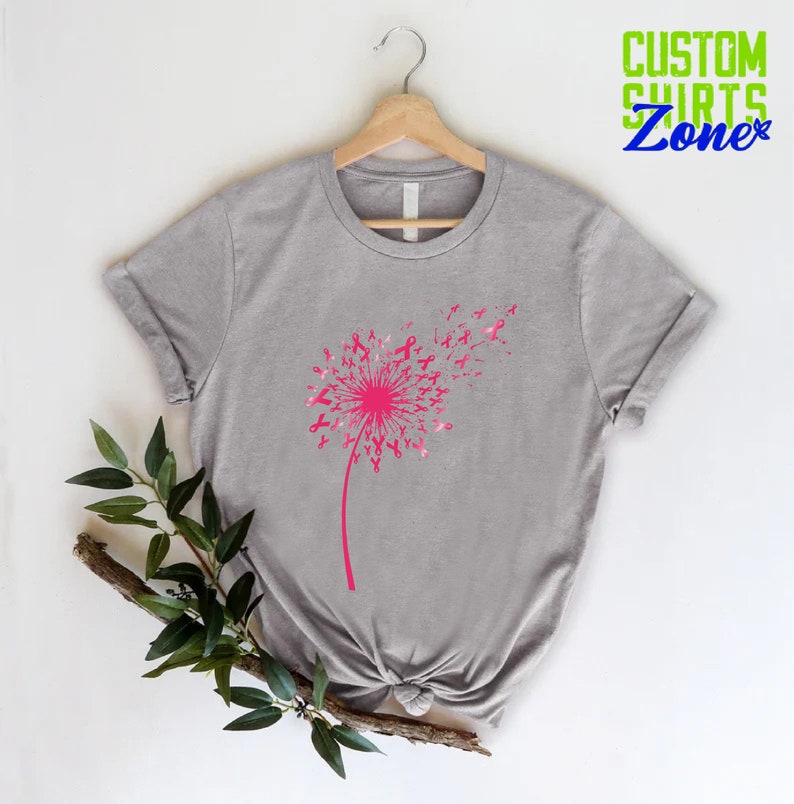Op de afbeelding: Grijs T-shirt met een roze paardenbloemontwerp gemaakt van roze linten. De paardenbloem waait in de wind. Het shirt hangt aan een hanger.