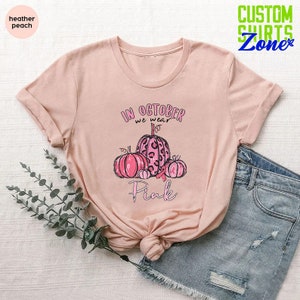 May include: A heather peach t-shirt with a pink graphic design featuring pumpkins and the text "In October we wear Pink".