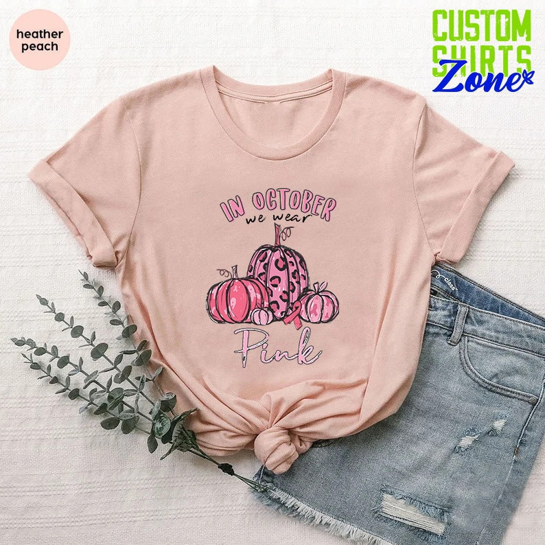 May include: A heather peach t-shirt with a pink graphic design featuring pumpkins and the text "In October we wear Pink".