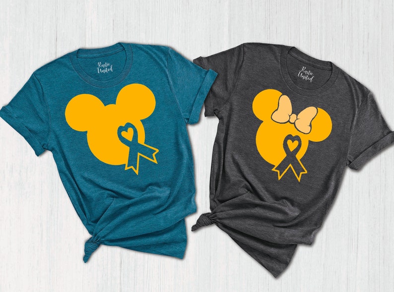 K&ouml;nnte beinhalten: Zwei T-Shirts mit einem gelben Mickey-Maus-Kopfdesign. Ein Shirt ist blaugr&uuml;n und das andere dunkelgrau. Beide Shirts haben ein blaues Band mit einem Herzen in der Mitte.