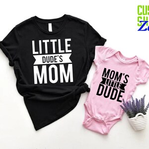Op de afbeelding: Een zwart t-shirt met de tekst "LITTLE DUDE'S MOM" en een roze babyromper met de tekst "MOM'S LITTLE DUDE". De afbeelding bevat ook een kleine witte pot met lavendel en de tekst "CUSTOM SHIRTS Zone".