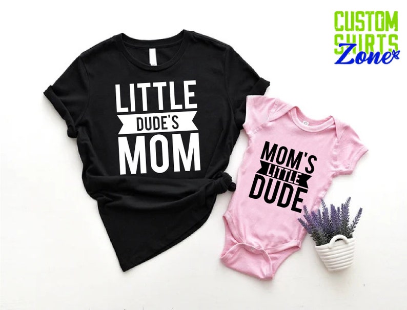 Op de afbeelding: Een zwart t-shirt met de tekst "LITTLE DUDE'S MOM" en een roze babyromper met de tekst "MOM'S LITTLE DUDE". De afbeelding bevat ook een kleine witte pot met lavendel en de tekst "CUSTOM SHIRTS Zone".