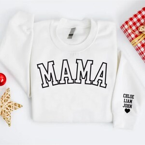 Puede incluir: Sudadera blanca con la palabra "MAMA" en letras negras de bloque en la parte delantera. La manga tiene los nombres "CHLOE, LIAM, JOHN" y un peque&ntilde;o coraz&oacute;n negro.