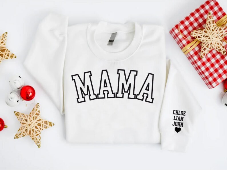 Puede incluir: Sudadera blanca con la palabra "MAMA" en letras negras de bloque en la parte delantera. La manga tiene los nombres "CHLOE, LIAM, JOHN" y un peque&ntilde;o coraz&oacute;n negro.