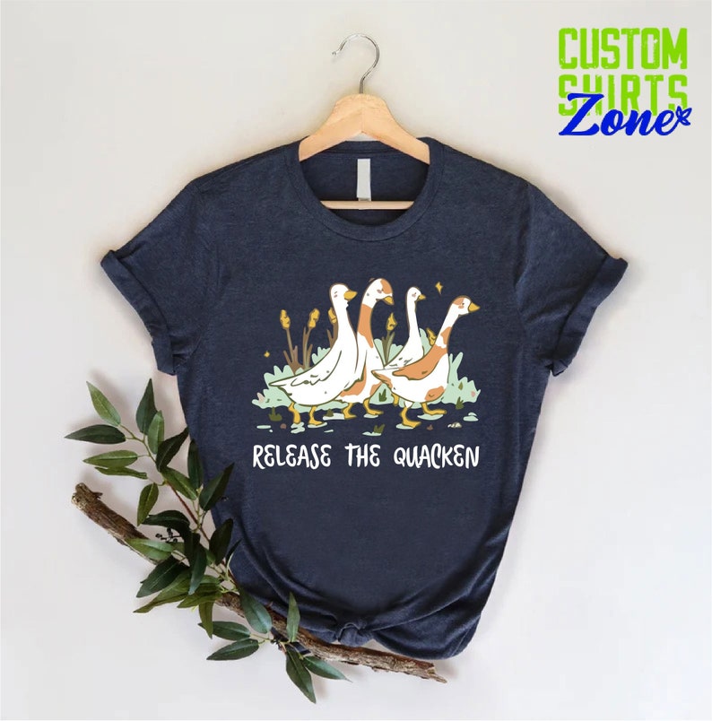 Pu&ograve; includere: Una maglietta blu navy con una grafica di quattro oche che camminano in fila. Il testo "Release the Quacken" &egrave; stampato sotto le oche.