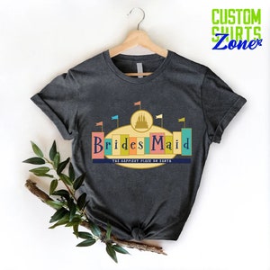 Puede incluir: Una camiseta gris oscuro con un gr&aacute;fico de un castillo y el texto "Brides Maid" en un dise&ntilde;o retro y colorido. El texto "The Happiest Place on Earth" tambi&eacute;n se incluye debajo del gr&aacute;fico.