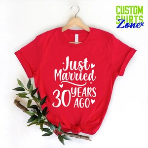 May include: A red t-shirt with the text "Just Married 30 Years Ago" in white script. The shirt is hanging on a wooden hanger. Green leaves and a branch are placed on the left side of the shirt. The words "Custom Shirts Zone" are in the upper right corner.