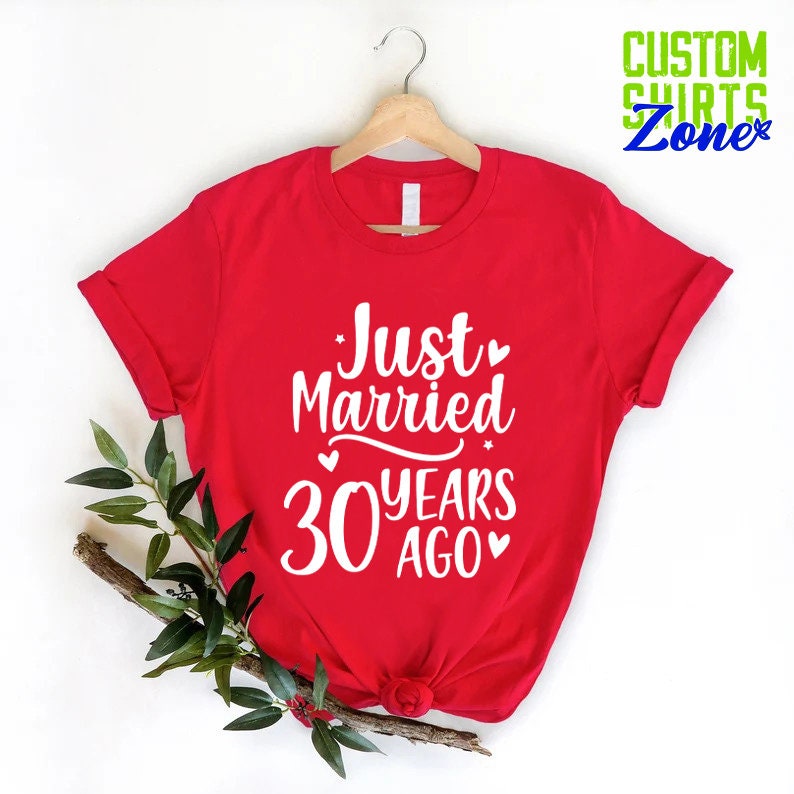 May include: A red t-shirt with the text "Just Married 30 Years Ago" in white script. The shirt is hanging on a wooden hanger. Green leaves and a branch are placed on the left side of the shirt. The words "Custom Shirts Zone" are in the upper right corner.