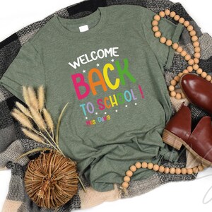 以下が含まれることがあります： カラフルな文字で「WELCOME BACK TO SCHOOL！」と書かれたオリーブグリーンのTシャツ。茶色の靴、木製のビーズネックレス、装飾的な織りのバスケットと組み合わせています。