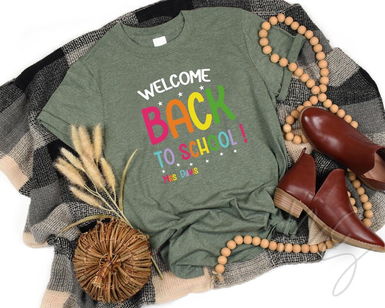 以下が含まれることがあります： カラフルな文字で「WELCOME BACK TO SCHOOL！」と書かれたオリーブグリーンのTシャツ。茶色の靴、木製のビーズネックレス、装飾的な織りのバスケットと組み合わせています。
