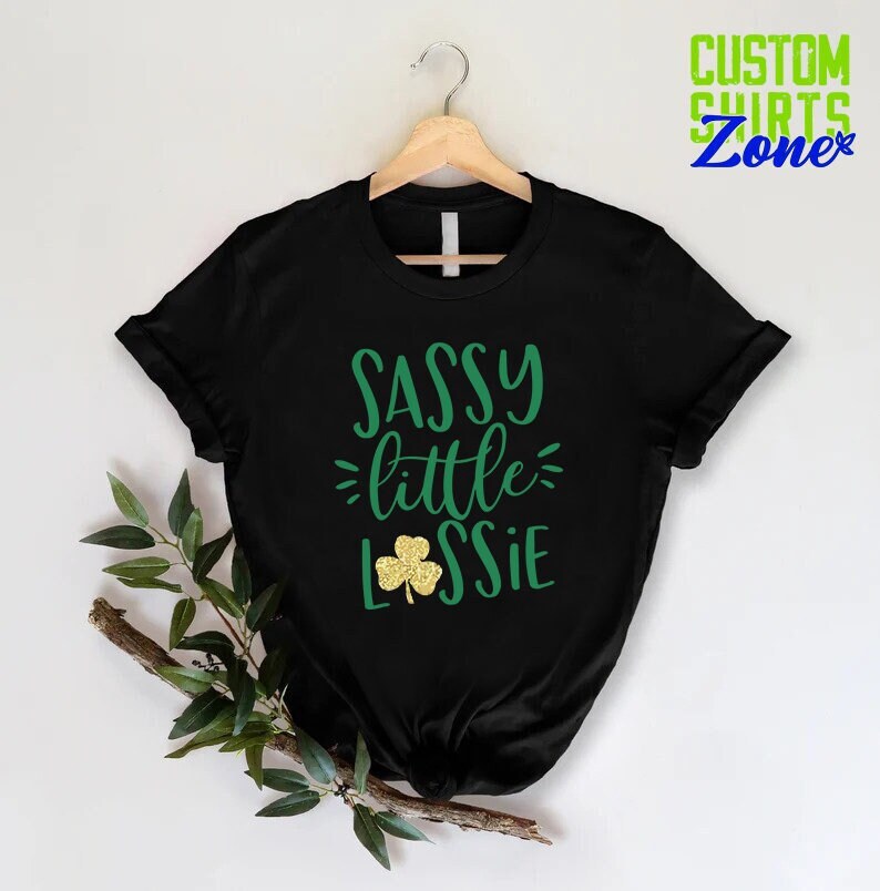 K&ouml;nnte beinhalten: Schwarzes T-Shirt mit gr&uuml;nem Text, der "Sassy little Lassie" lautet, mit einem goldenen Glitzer-Kleeblatt.