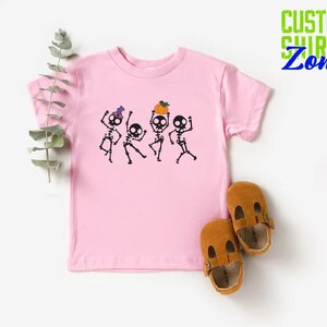 May include: Pink t-shirt with a graphic of four skeletons dancing. The skeletons are wearing hats and holding pumpkins. The shirt is a toddler size.