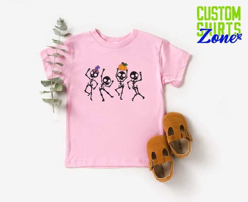 May include: Pink t-shirt with a graphic of four skeletons dancing. The skeletons are wearing hats and holding pumpkins. The shirt is a toddler size.