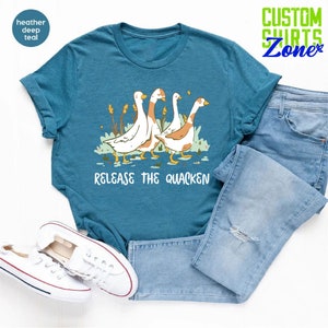 May include: A teal blue t-shirt with a graphic of four geese walking in a line. The geese are white with brown heads and necks. The text "Release the Quacken" is printed below the geese.