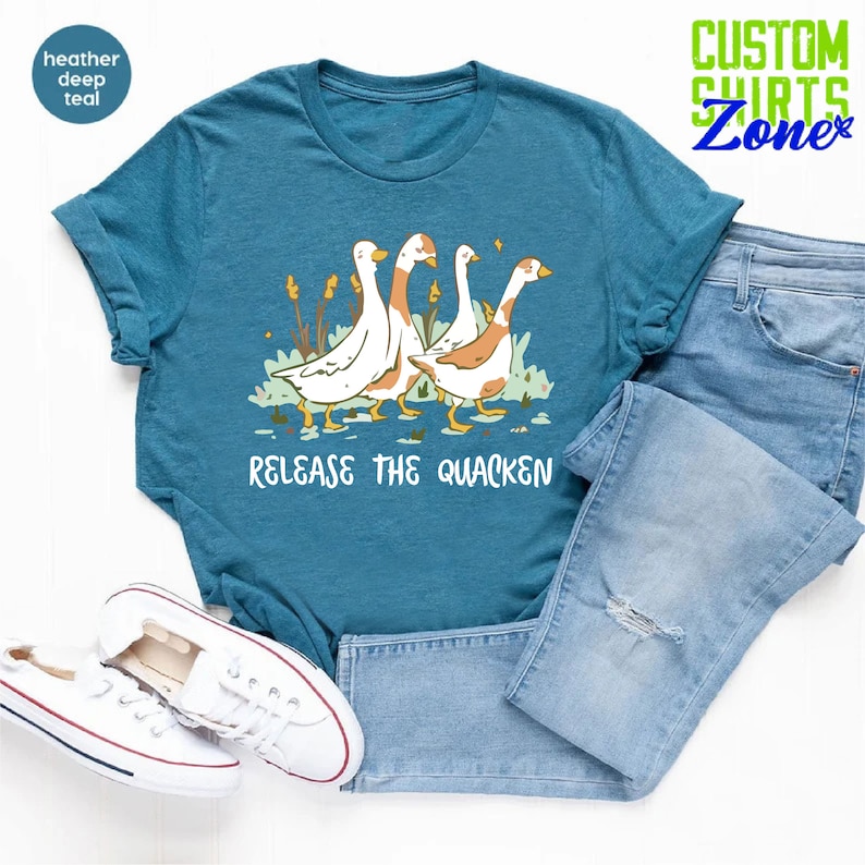 May include: A teal blue t-shirt with a graphic of four geese walking in a line. The geese are white with brown heads and necks. The text "Release the Quacken" is printed below the geese.
