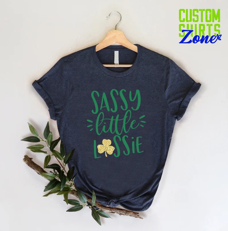K&ouml;nnte beinhalten: Ein dunkelblaues T-Shirt mit dem Text "SASSY little LSSIE" in gr&uuml;ner Schrift. Unter dem Text befindet sich ein goldener Glitzer-Kleeblatt.