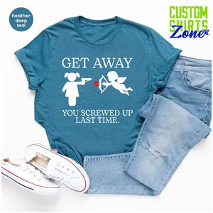 May include: A teal blue t-shirt with the text "GET AWAY YOU SCREWED UP LAST TIME."  The text is in white and there is a white illustration of a woman pointing a gun at a cupid with an arrow.