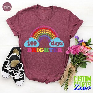 以下が含まれることがあります： ヘザーマロンのTシャツに、ハートでできた虹と、「100日輝き続ける」というカラフルな文字が書かれています。