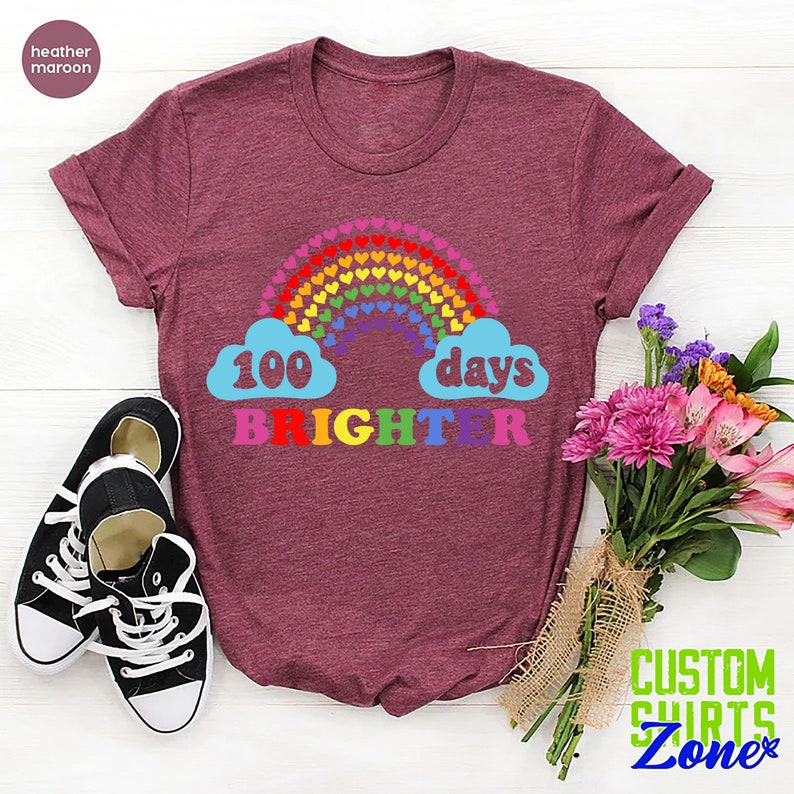 以下が含まれることがあります： ヘザーマロンのTシャツに、ハートでできた虹と、「100日輝き続ける」というカラフルな文字が書かれています。